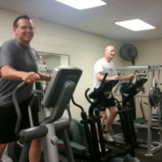 Two men in a gym using elliptical machines